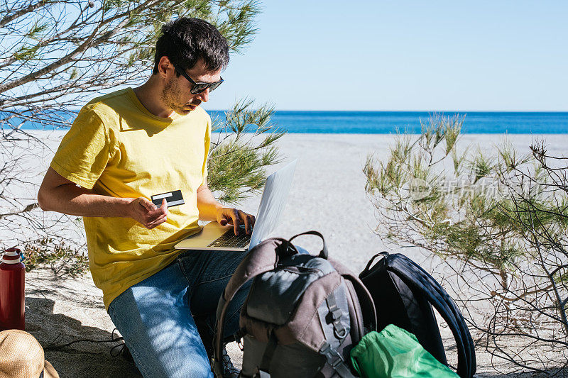 数码游民(Digital nomad traveler)在海边的海滩上用笔记本电脑用信用卡在线购物。